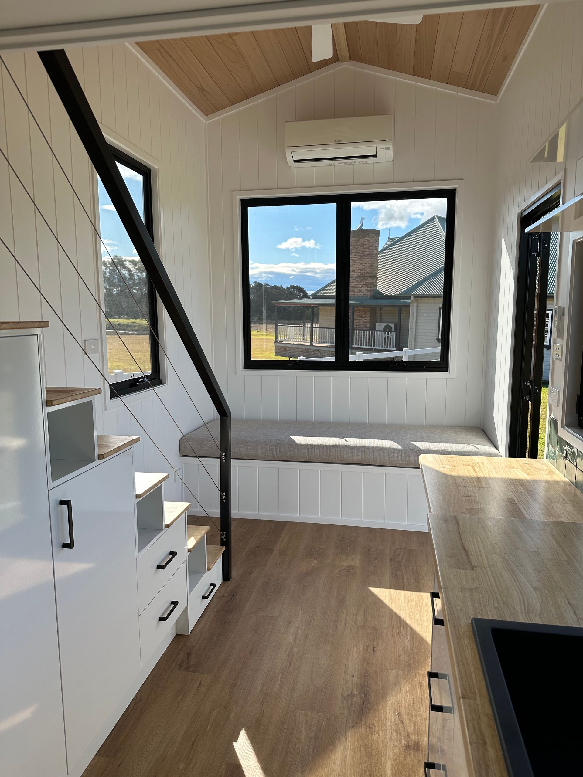 Tiny house interior with stairs, bench seating, and a large window overlooking a yard.