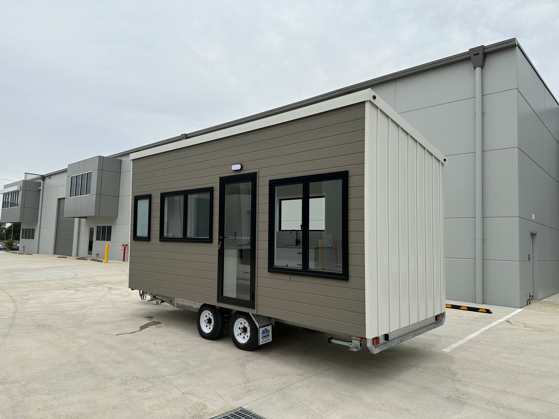 Tiny house trailer with brown and white exterior, parked near a gray industrial building.