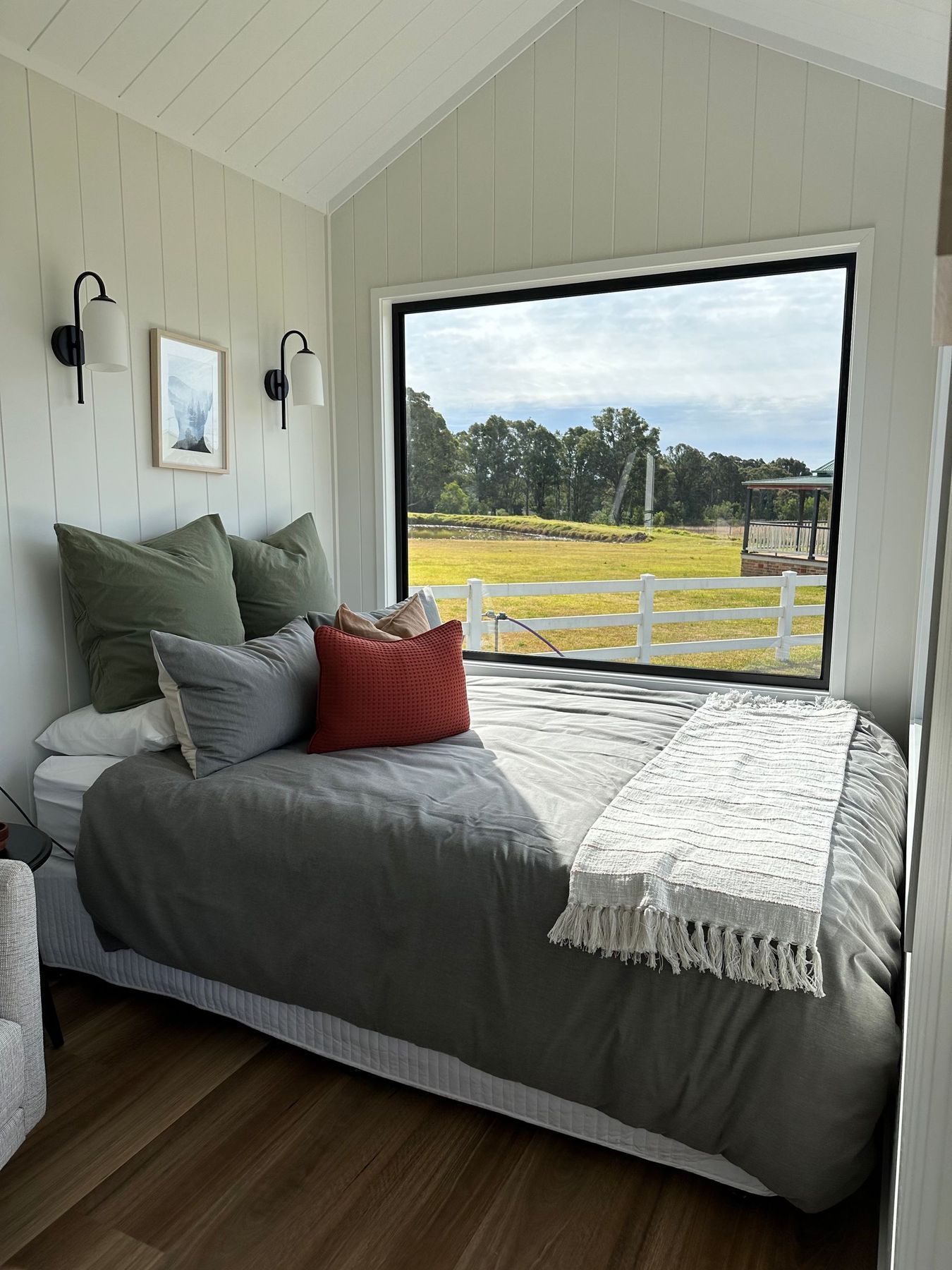 Cozy bedroom with twin bed, green/red pillows, large window overlooking a field, white walls, and black sconces.