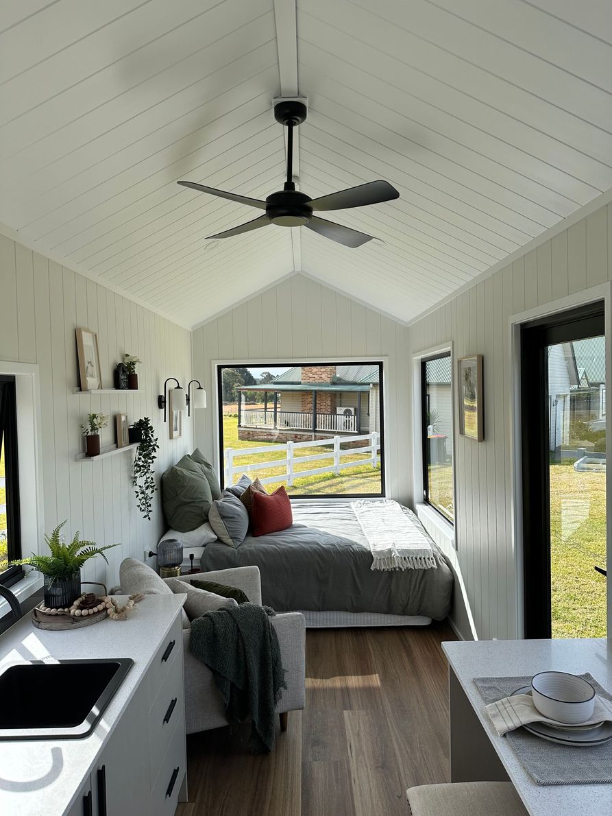 Tiny wooden house on wheels with black window and trailer hitch on a grassy field.
