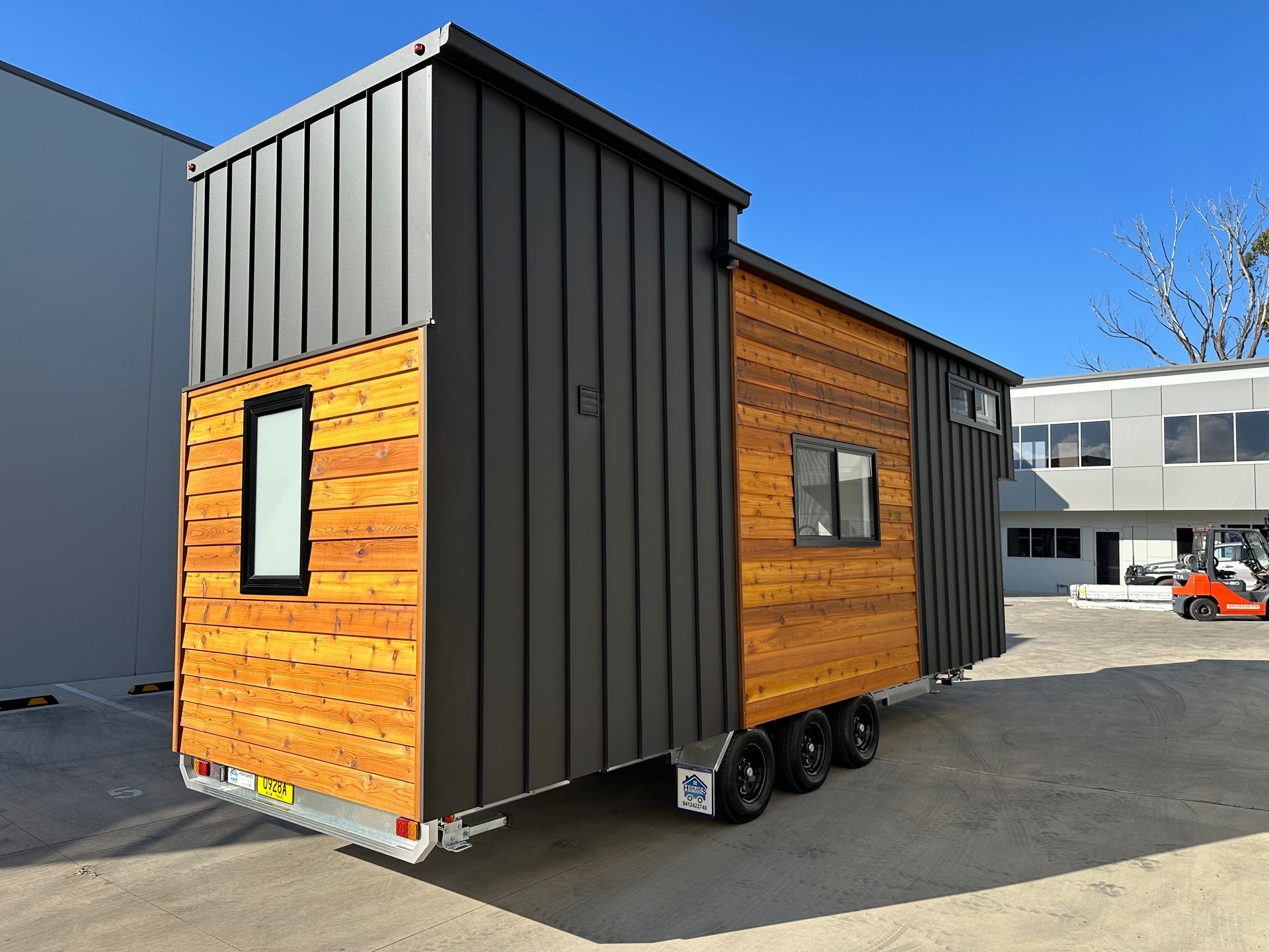 Tiny house trailer with a wood-paneled side and black metal roof and accents, on a sunny day.