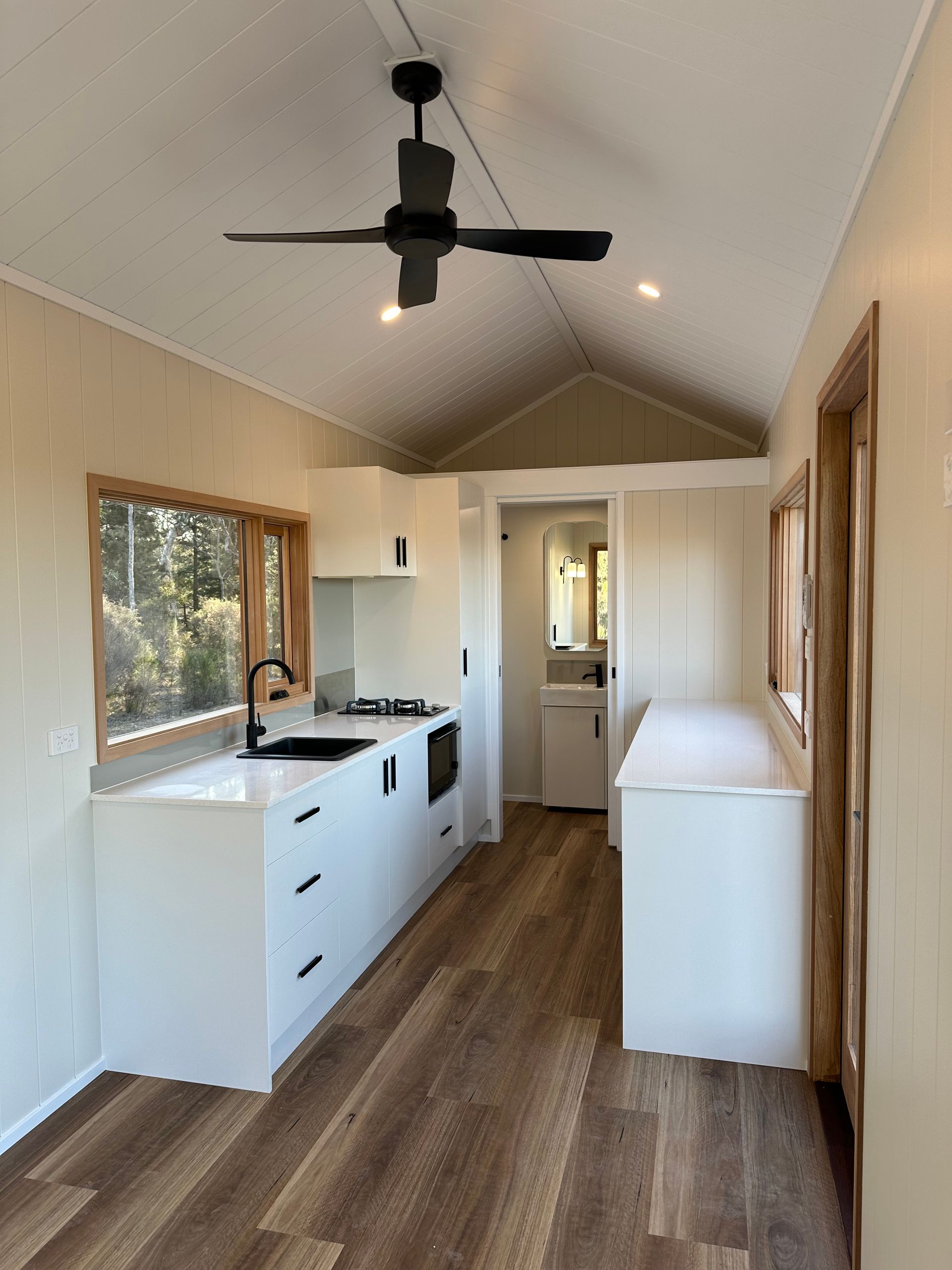 A tiny kitchen with white cabinets, a black stovetop, and a ceiling fan, in a narrow, wood-floored space.