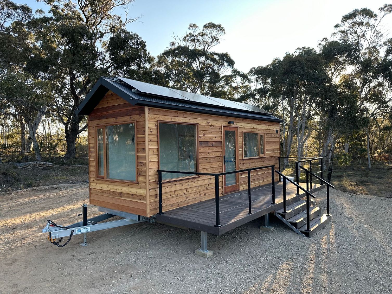 Tiny wooden house on wheels with solar panels, deck, and steps; outdoors.