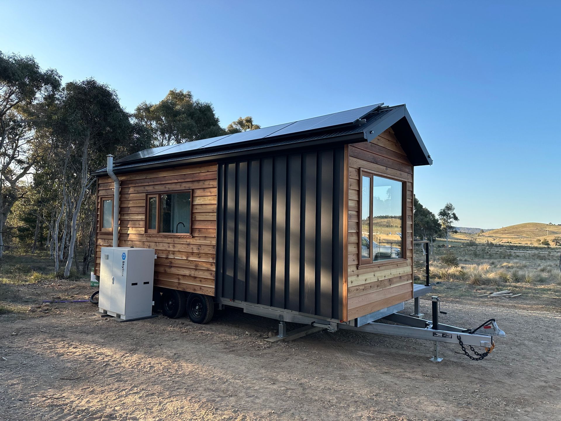 Tiny wooden cabin on wheels with solar panels, parked on gravel in a natural setting.