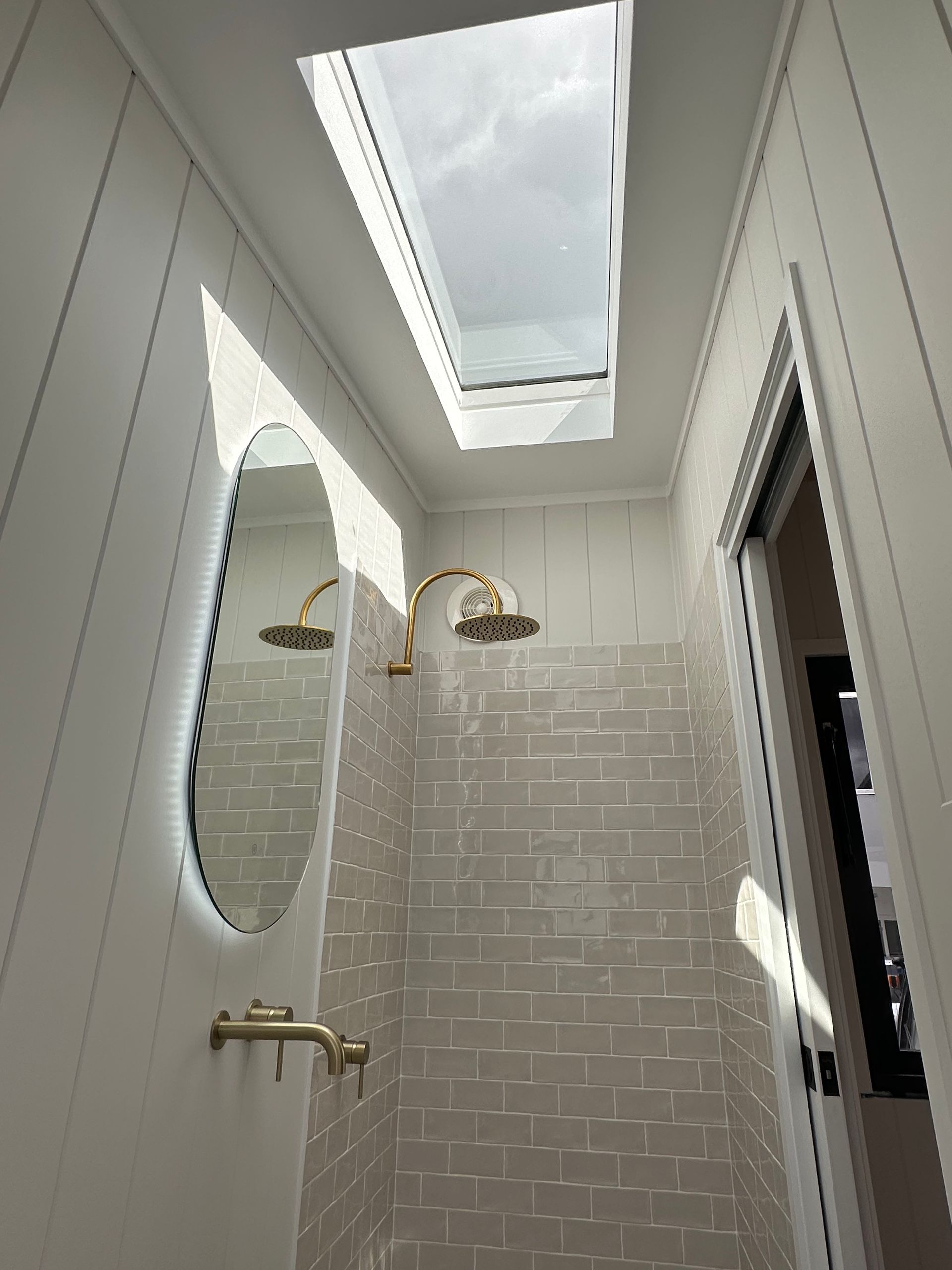 Narrow bathroom with skylight, gold fixtures, and light gray brick tile. Oval mirror on the left.