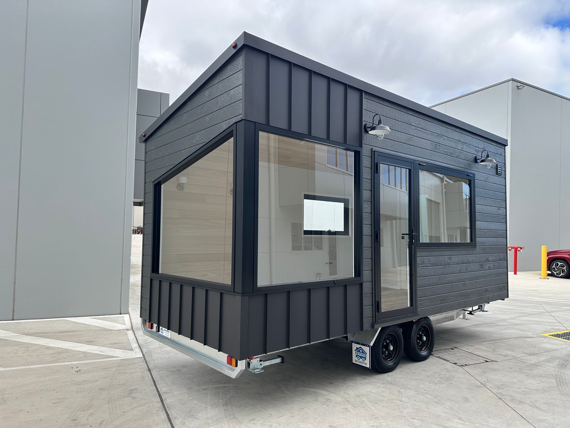 A modern, dark gray tiny house on wheels with large windows, parked in a concrete lot.