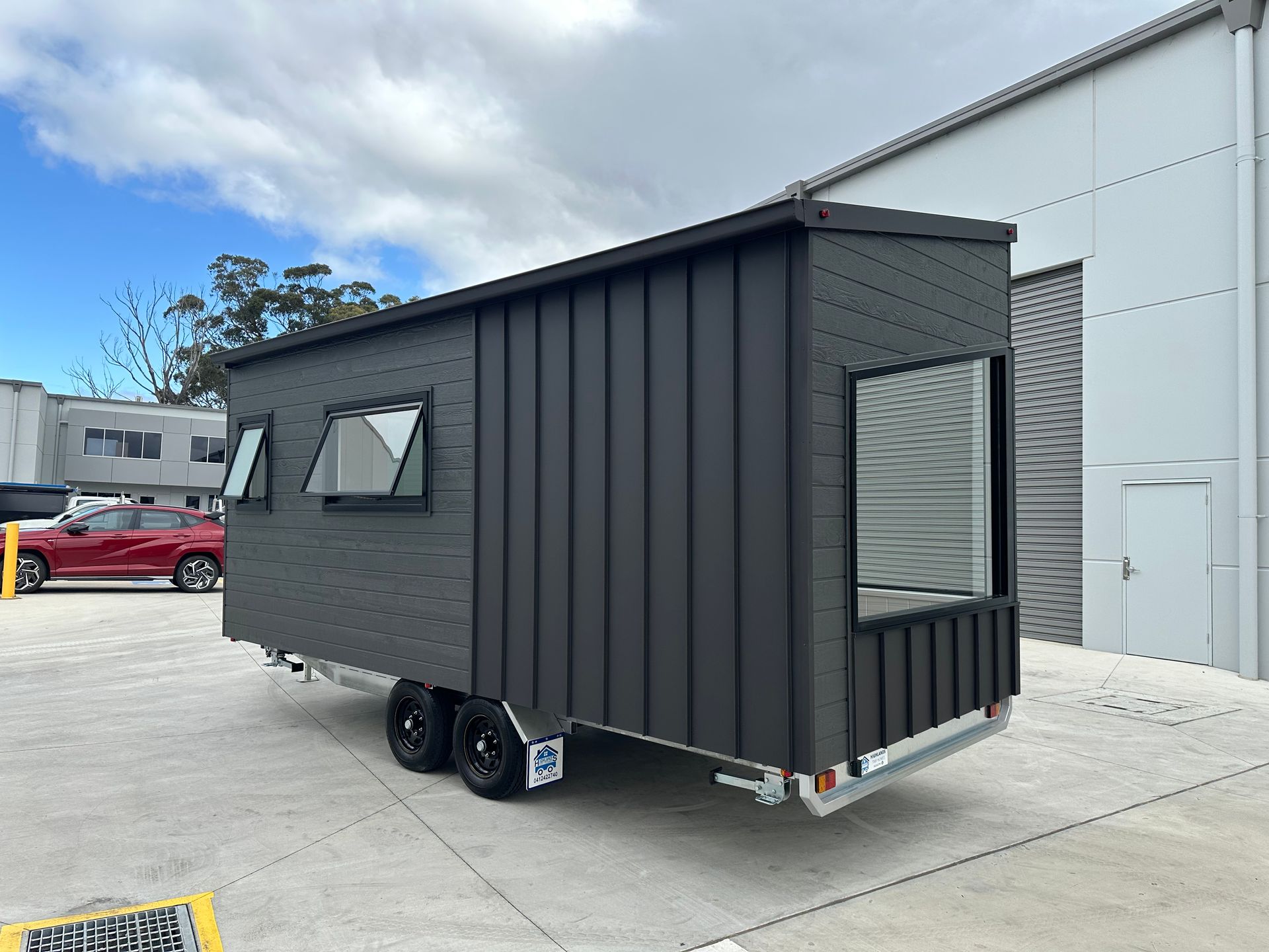 Dark gray tiny house on wheels, with windows, parked in front of a building and a red car.