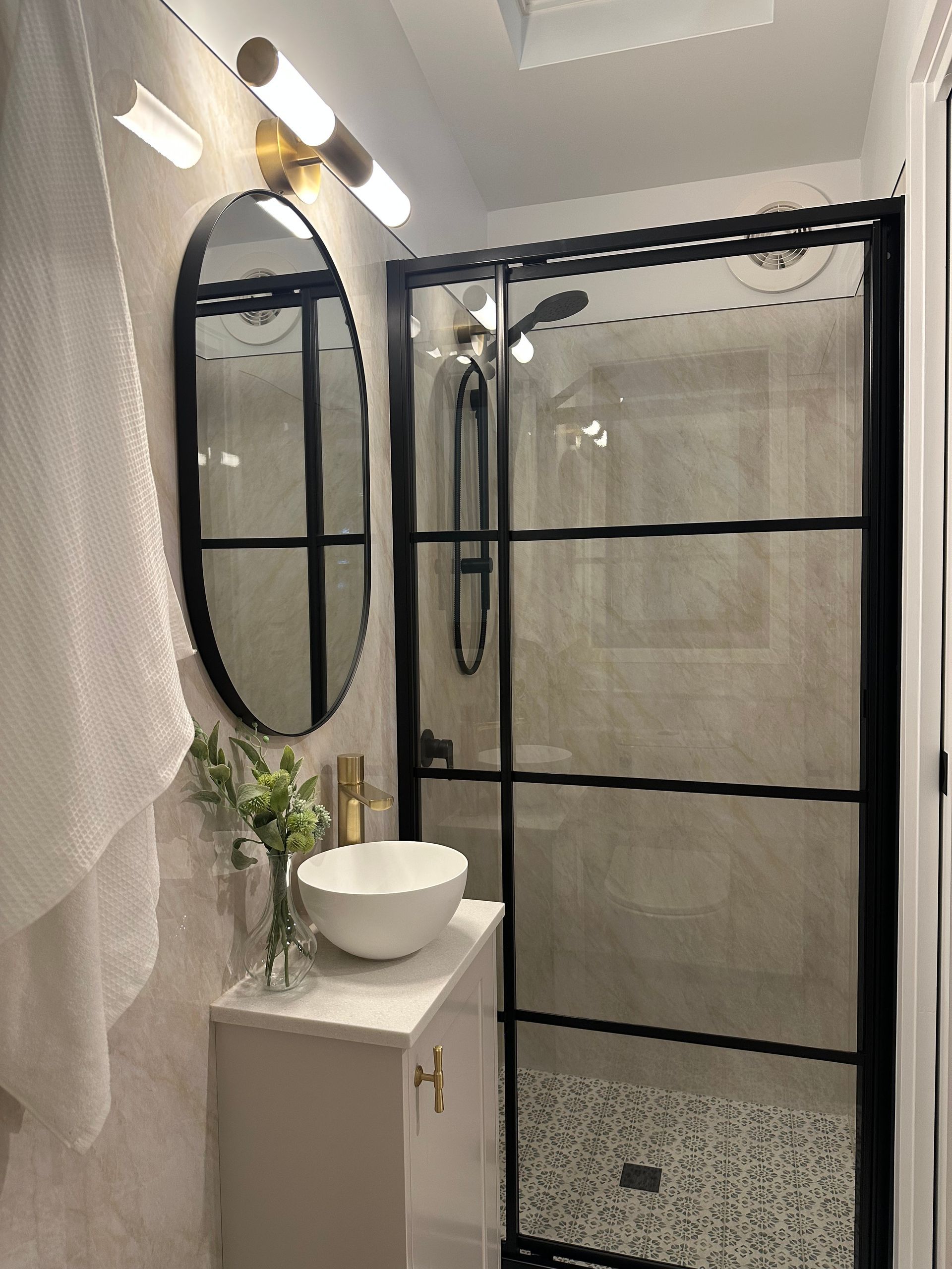 Modern bathroom: oval mirror, black-framed shower, white sink on cabinet, neutral tile, and brass accents.