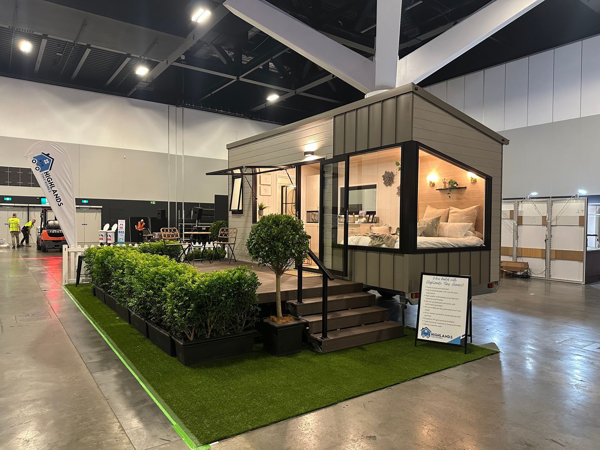 Cozy, bright tiny home interior with bed by large window, white cabinets, and wood floors.