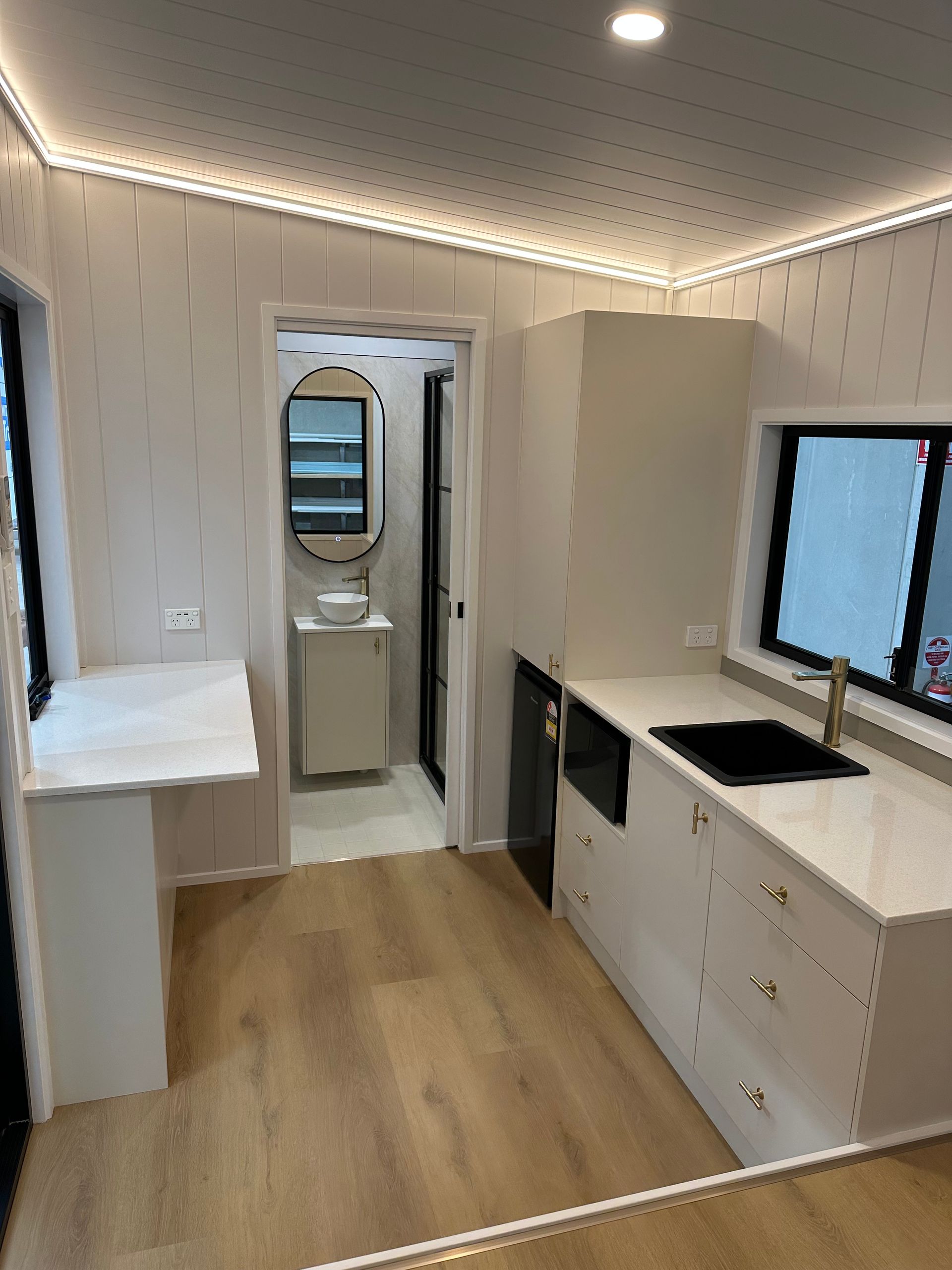 Interior view of a modern tiny home with kitchen, bathroom, light-colored cabinetry, and wood flooring.