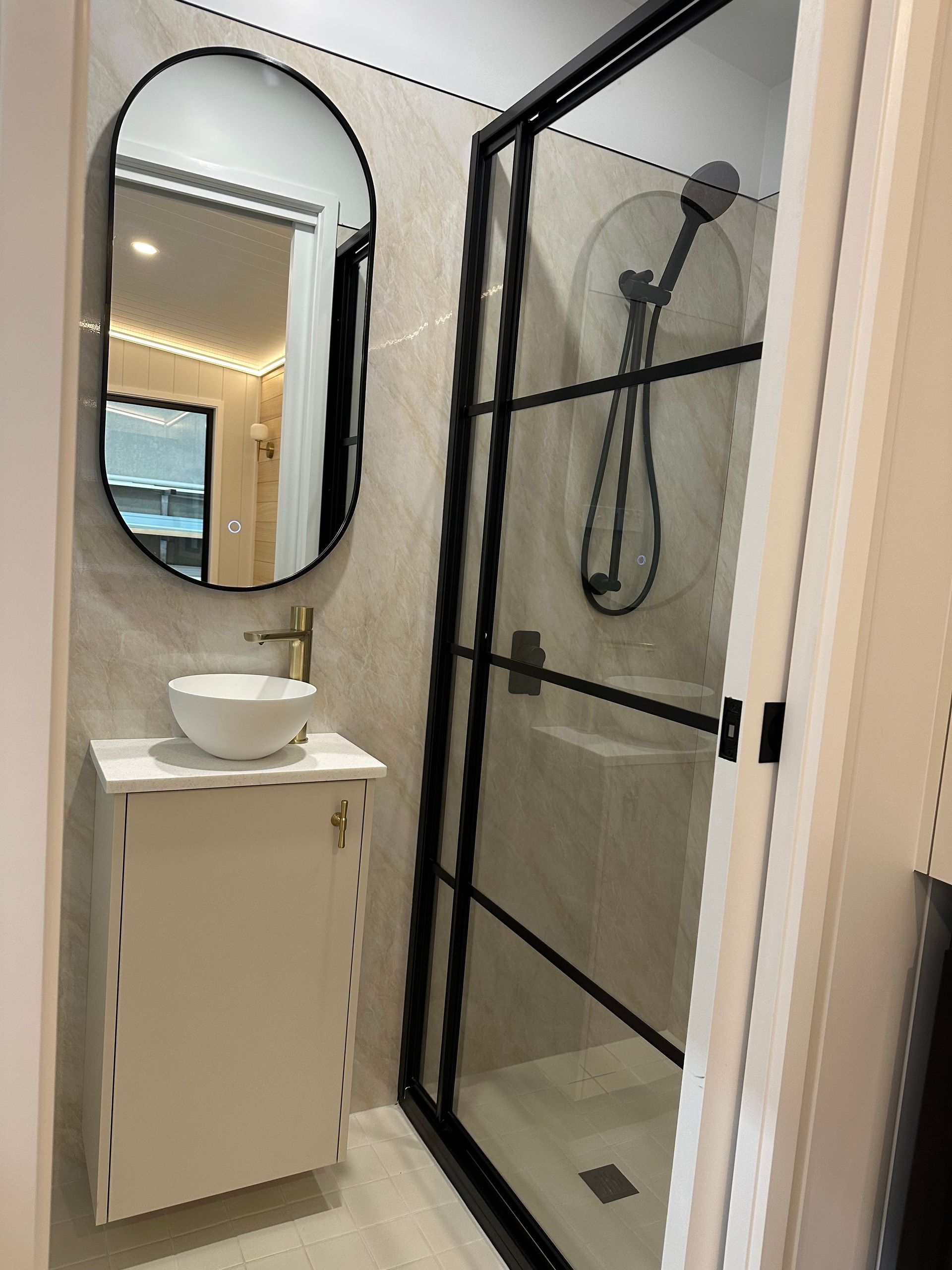 Small modern bathroom with shower, vanity, oval mirror. Black shower door frame, white sink, beige tile.