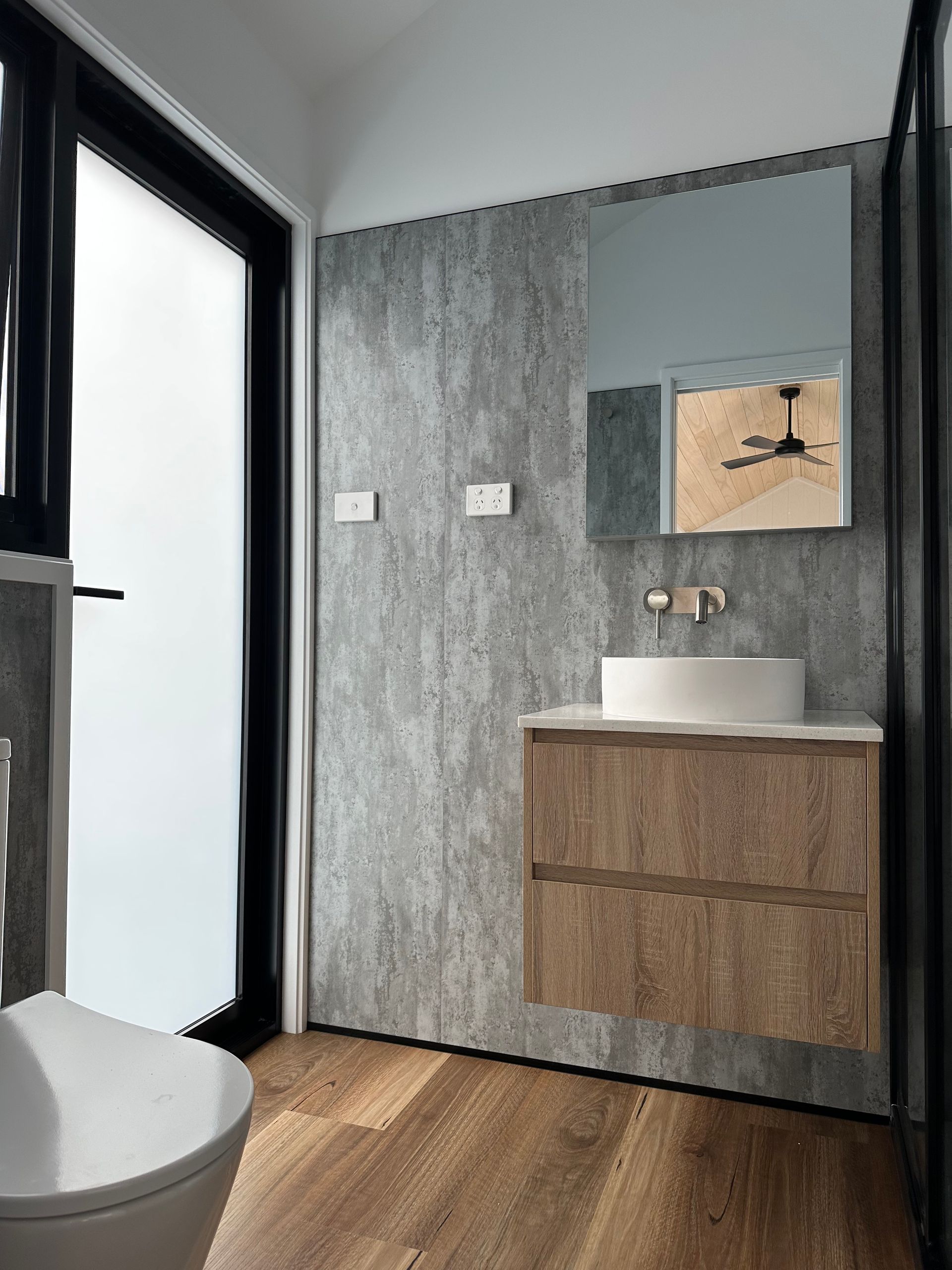 Bathroom with a wooden vanity, round sink, large mirror, and frosted window; gray textured wall and wooden floor.