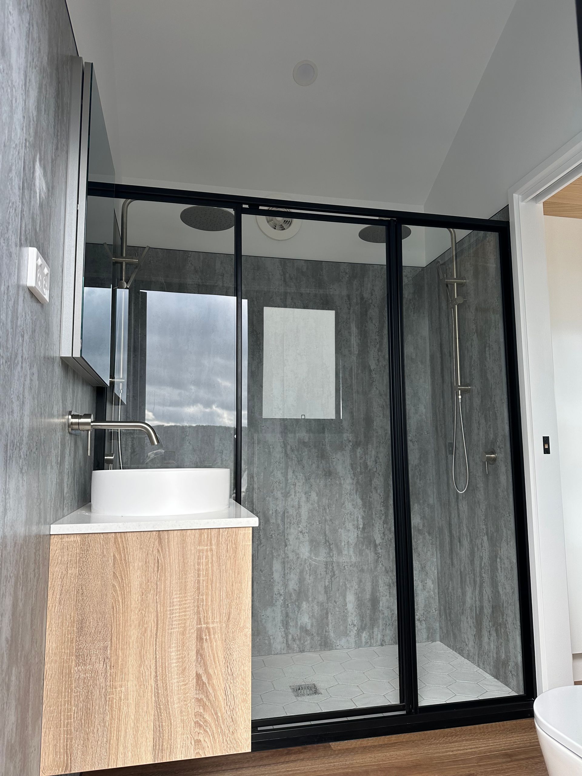 Modern bathroom with gray walls, wood vanity, glass shower with black frame.