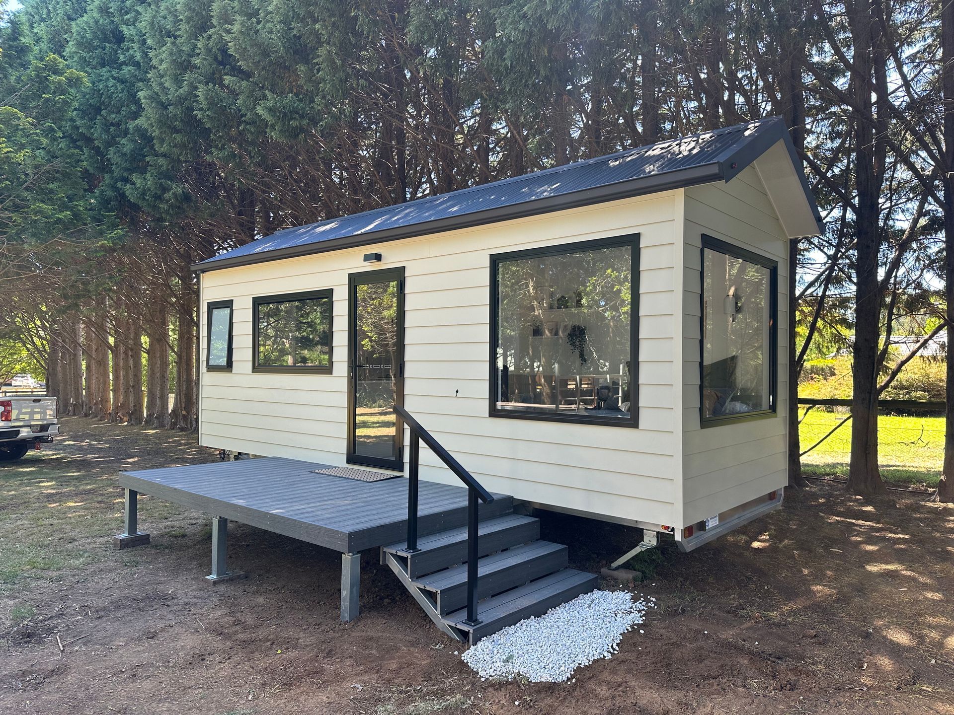 Small, light-colored tiny home with black trim and large windows, surrounded by trees.
