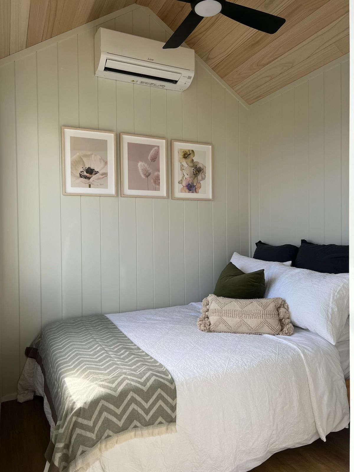 Bedroom with white bed, green blanket, three framed floral prints, and air conditioning unit.