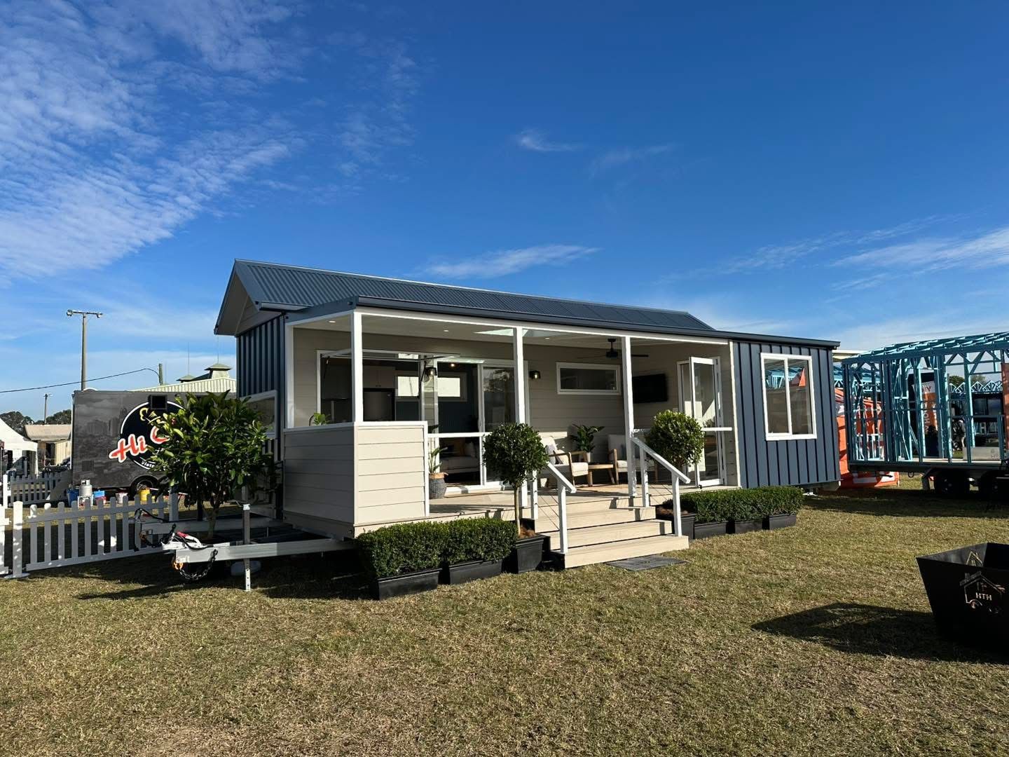 A blue and tan tiny home sits on a trailer on a grassy field under a blue sky.