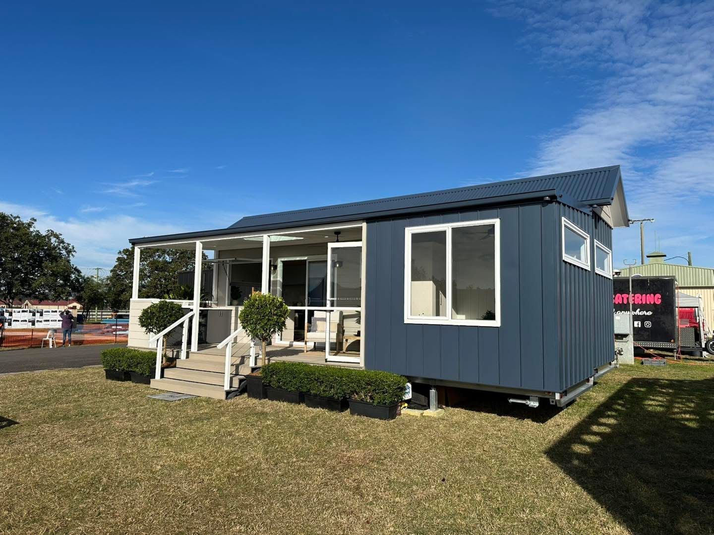 Blue tiny house with porch on grass under a blue sky.