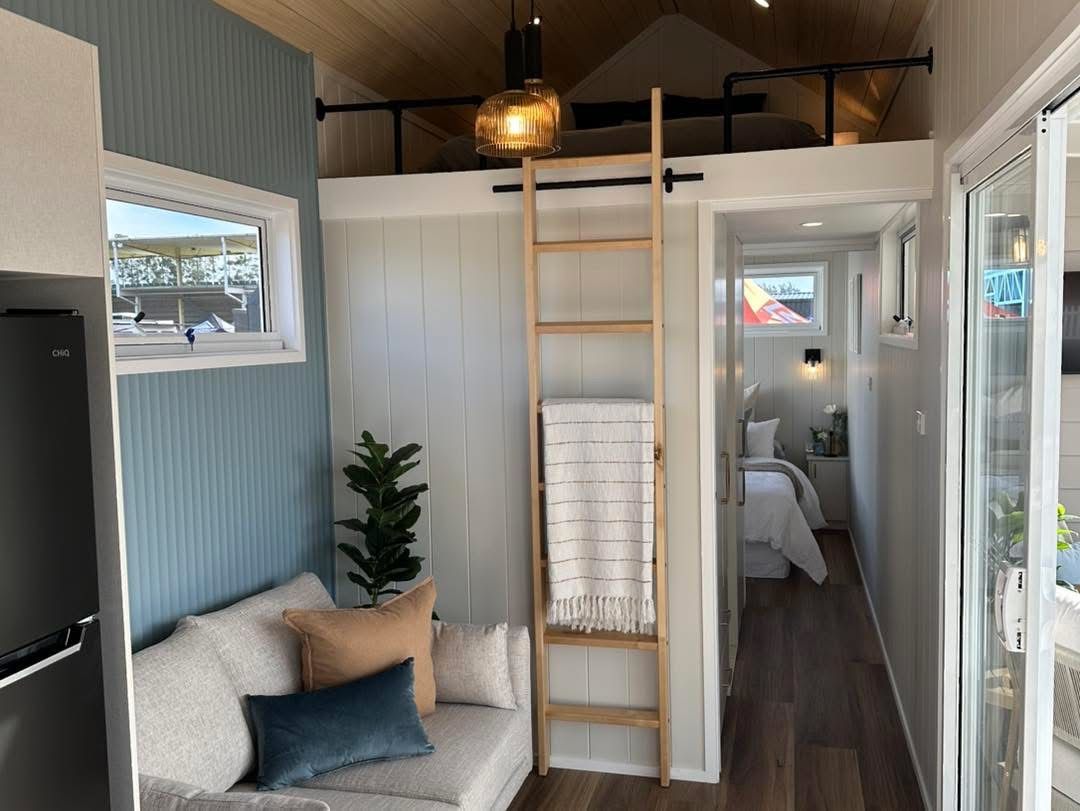 Tiny house interior with a loft, sofa, ladder, and bedroom visible through the doorway.
