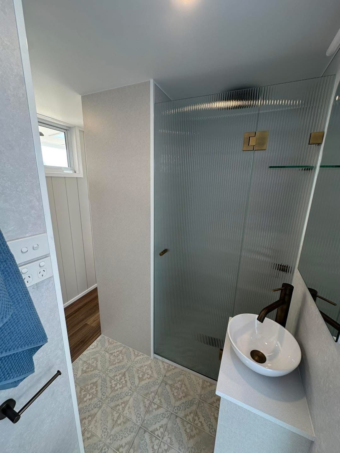 Small, modern bathroom with patterned floor, white sink, glass shower, and gold fixtures.