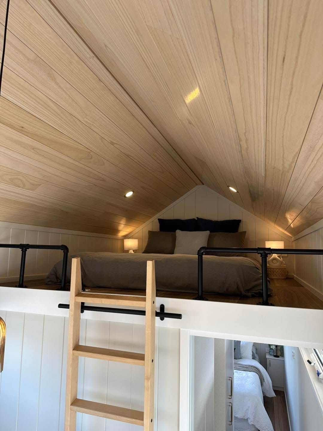 Loft bedroom in a tiny house with wooden ceiling, bed, ladder, and small lamps.