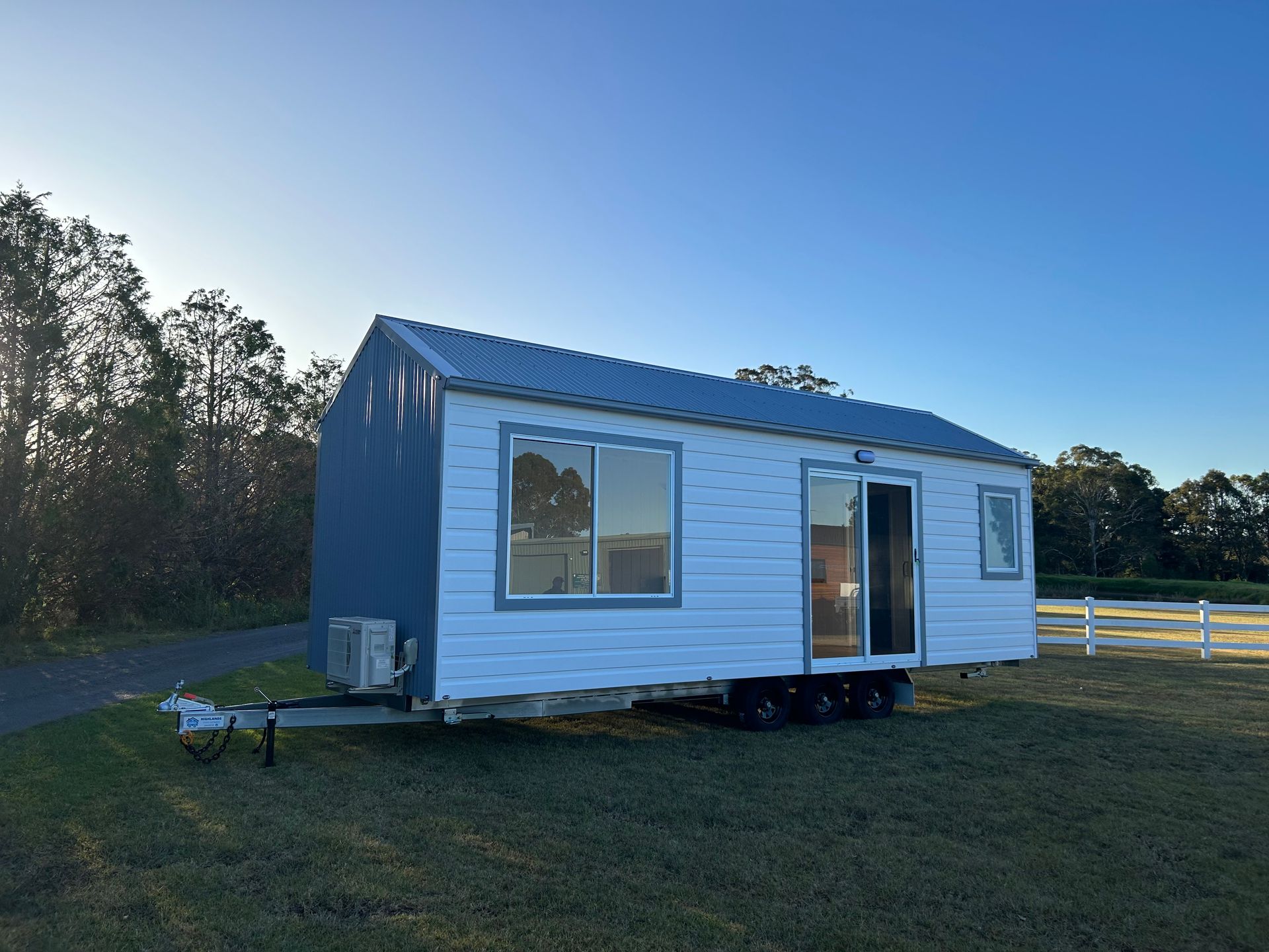 Tiny white house on wheels, blue roof, parked on grass. Bright daylight.