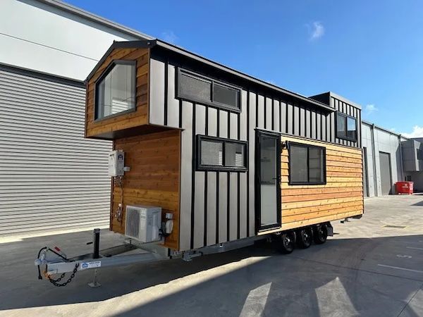 Tiny house on wheels with gray siding and wood accents, parked outside on a sunny day.