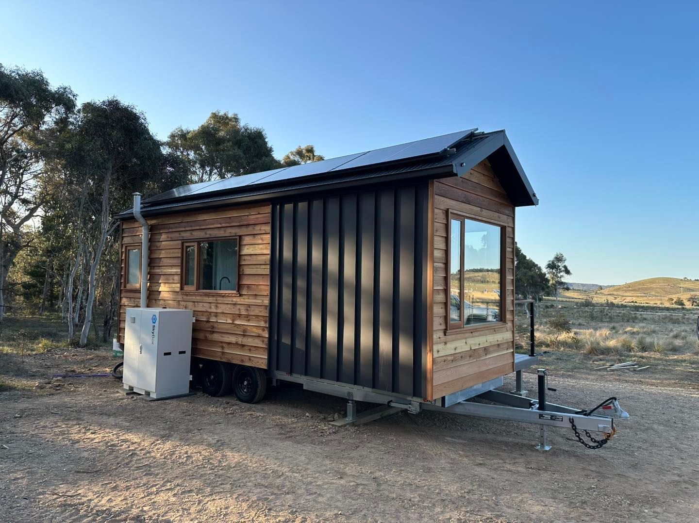 Tiny wooden house on wheels with black window and trailer hitch on a grassy field.