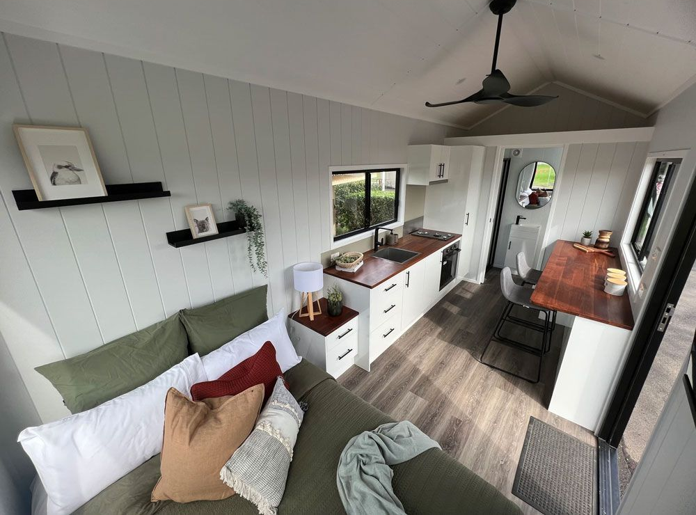 Tiny home interior with couch, kitchen, desk, and bathroom. White walls, wood accents, and a ceiling fan.