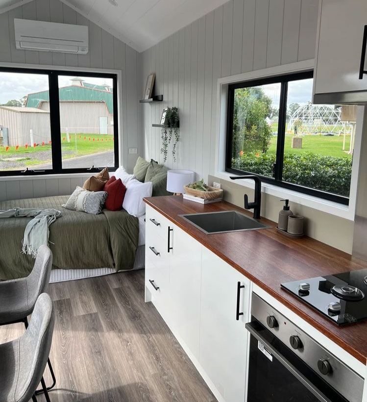 Tiny home interior: Kitchen with white cabinets, wood countertop, and a bed near windows.