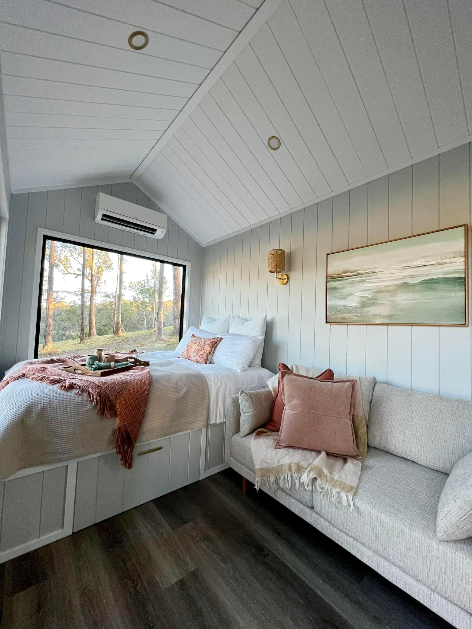 Cozy bedroom with bed, sofa, large window with a forest view, and wood floors.