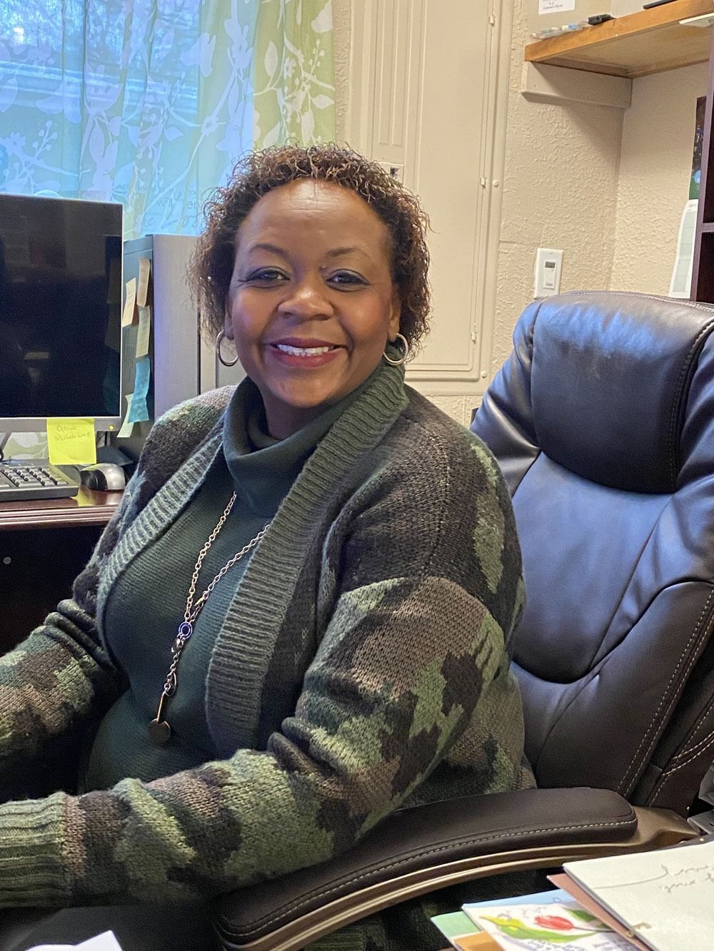 A woman is sitting in a chair in front of a computer and smiling.