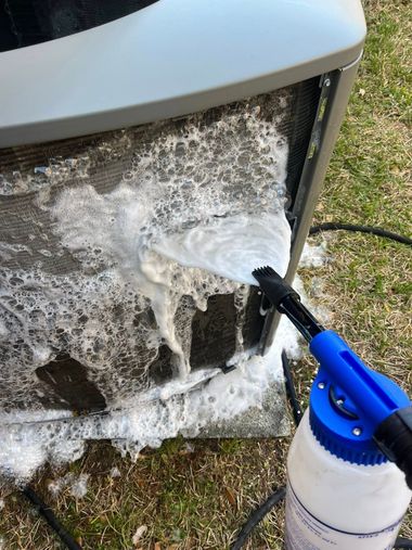 Pressure washer spraying foam and water onto a dirty outdoor appliance or unit on grass.