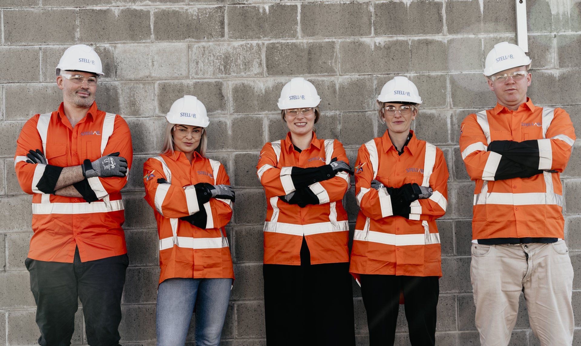Five construction workers, wearing orange vests and hard hats, arms crossed, standing in front of a gray brick wall.