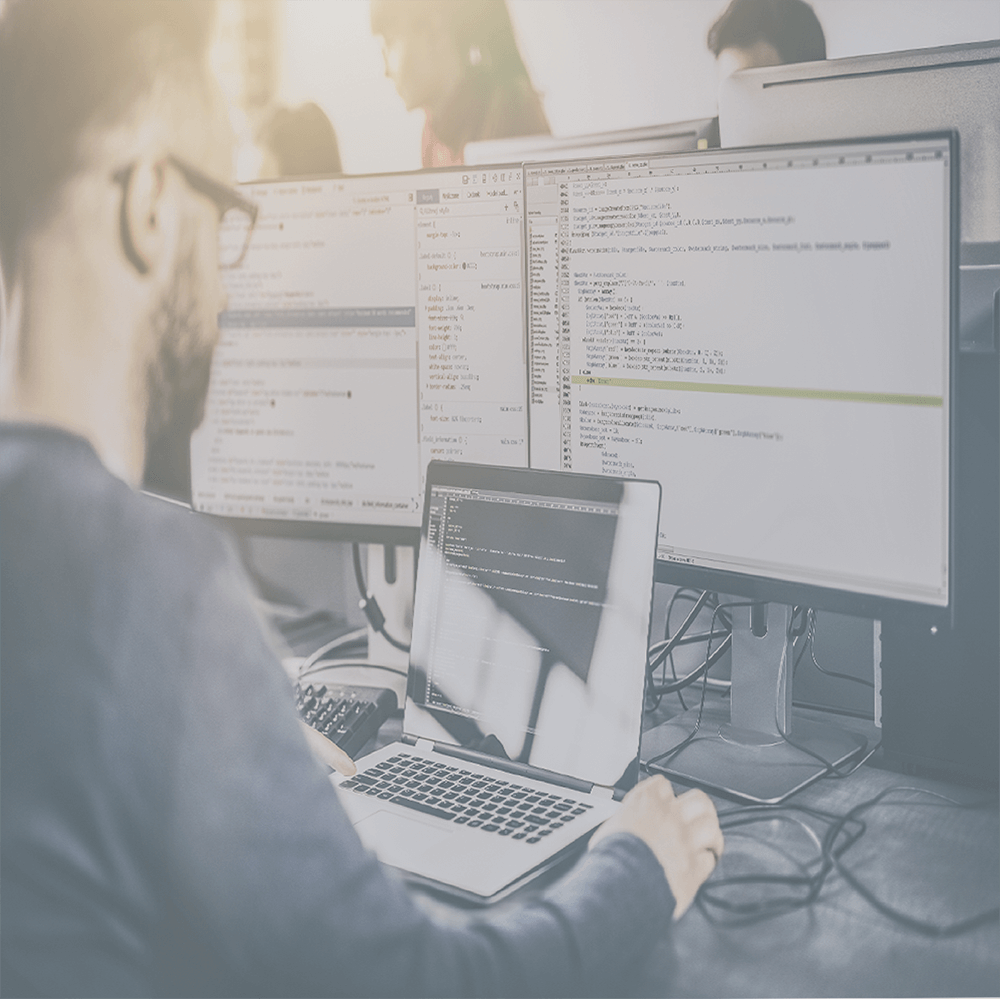 Person coding at a computer with two monitors, typing on laptop keyboard in an office.