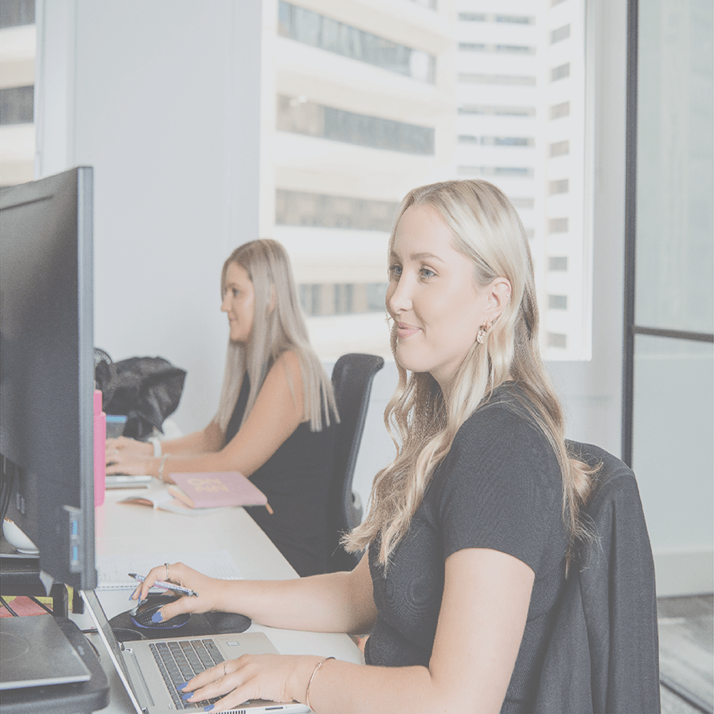 Two blonde women working at desks in an office, looking at computers.