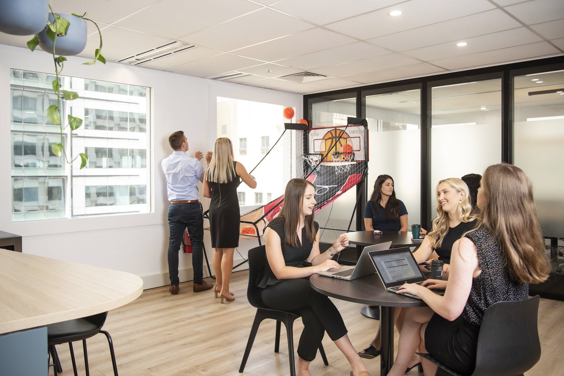 Office scene: people working at a table, others playing basketball, a bright office space.