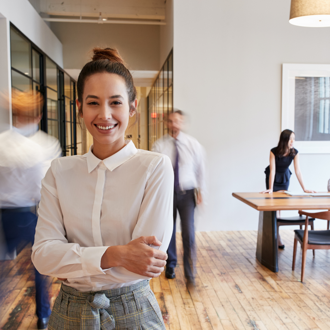 Woman in office smiling, arms crossed. Others blurred in background.