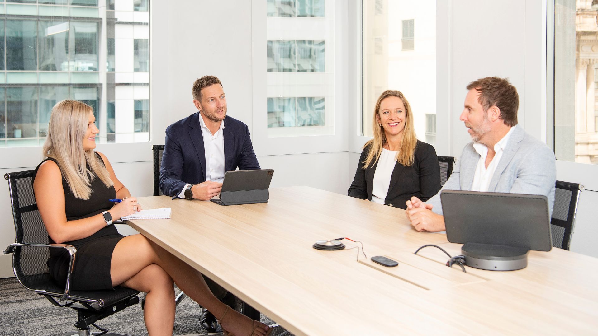 Four professionals in an office meeting, seated around a table, smiling, discussing ideas.