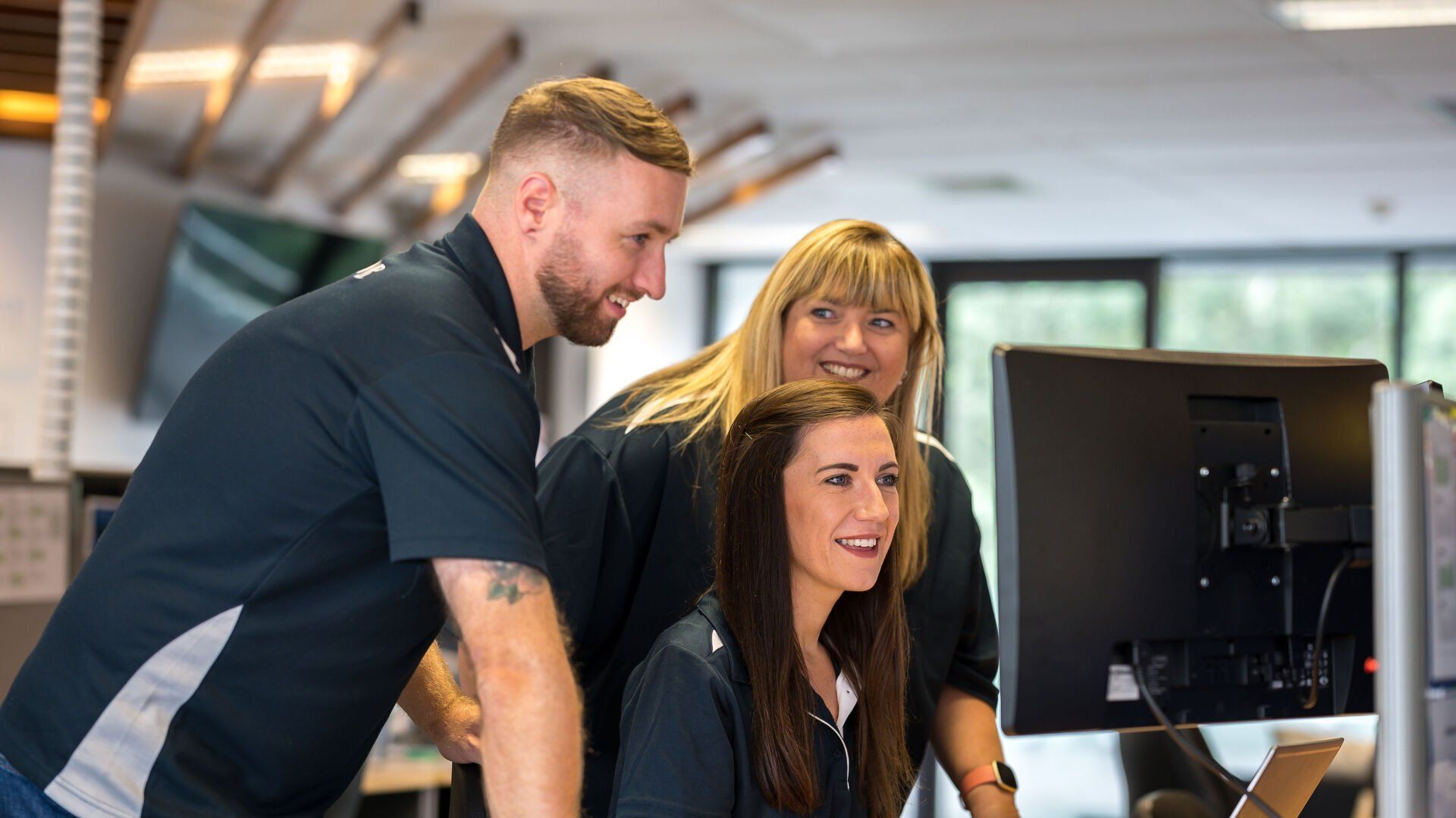 Three colleagues smiling, looking at a computer screen in an office.
