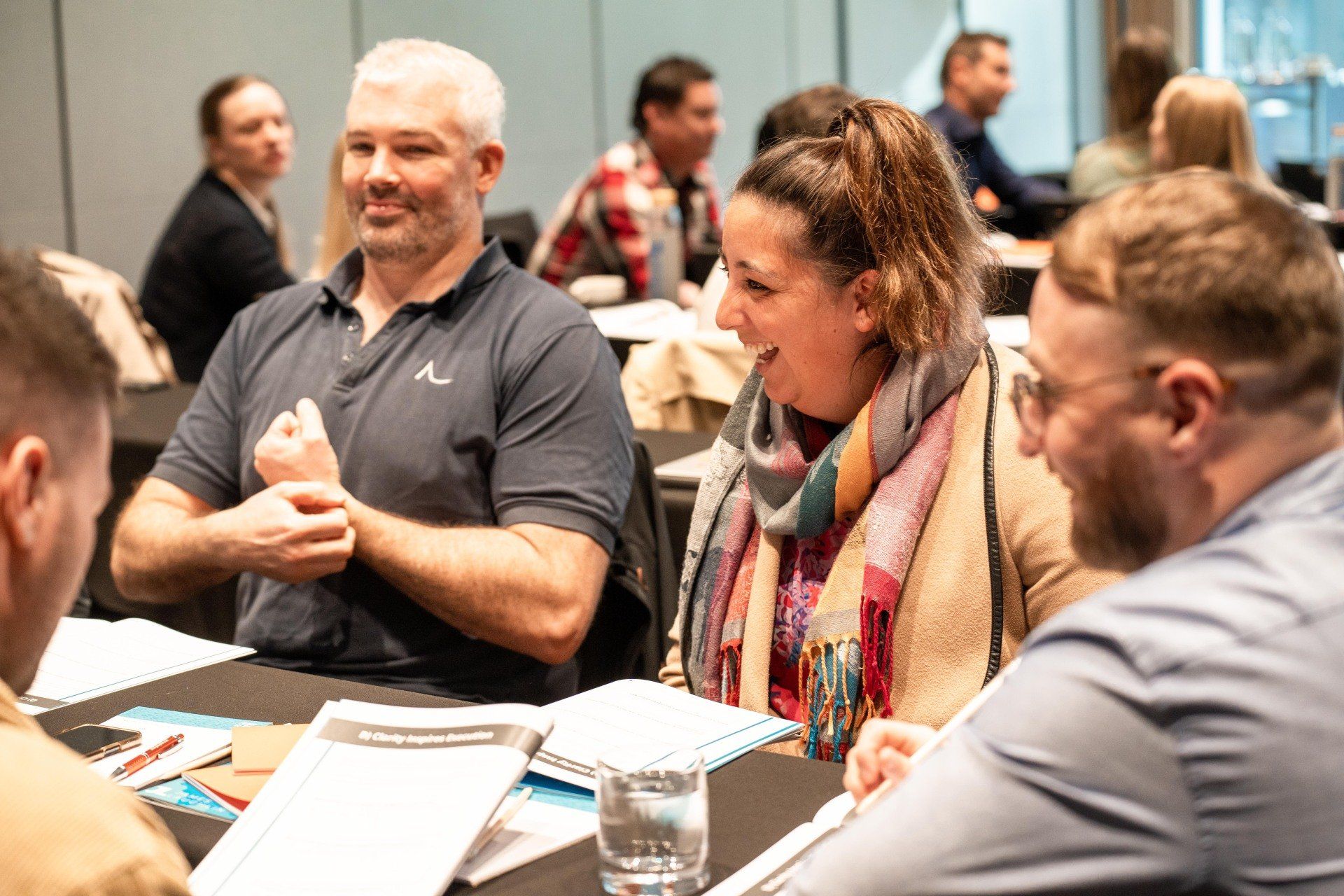 People at a meeting, laughing and gesturing. A woman in a scarf is smiling, and a man in grey shirt is gesturing.