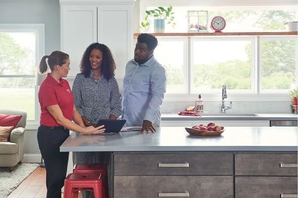 A woman in a red shirt shows a tablet to a couple in a bright kitchen.