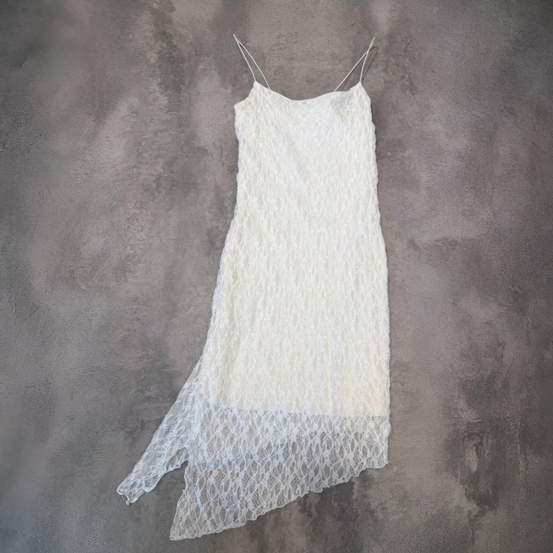 Vestido lencero de encaje blanco con tirantes finos y bajo asimétrico, colgado sobre una pared gris.