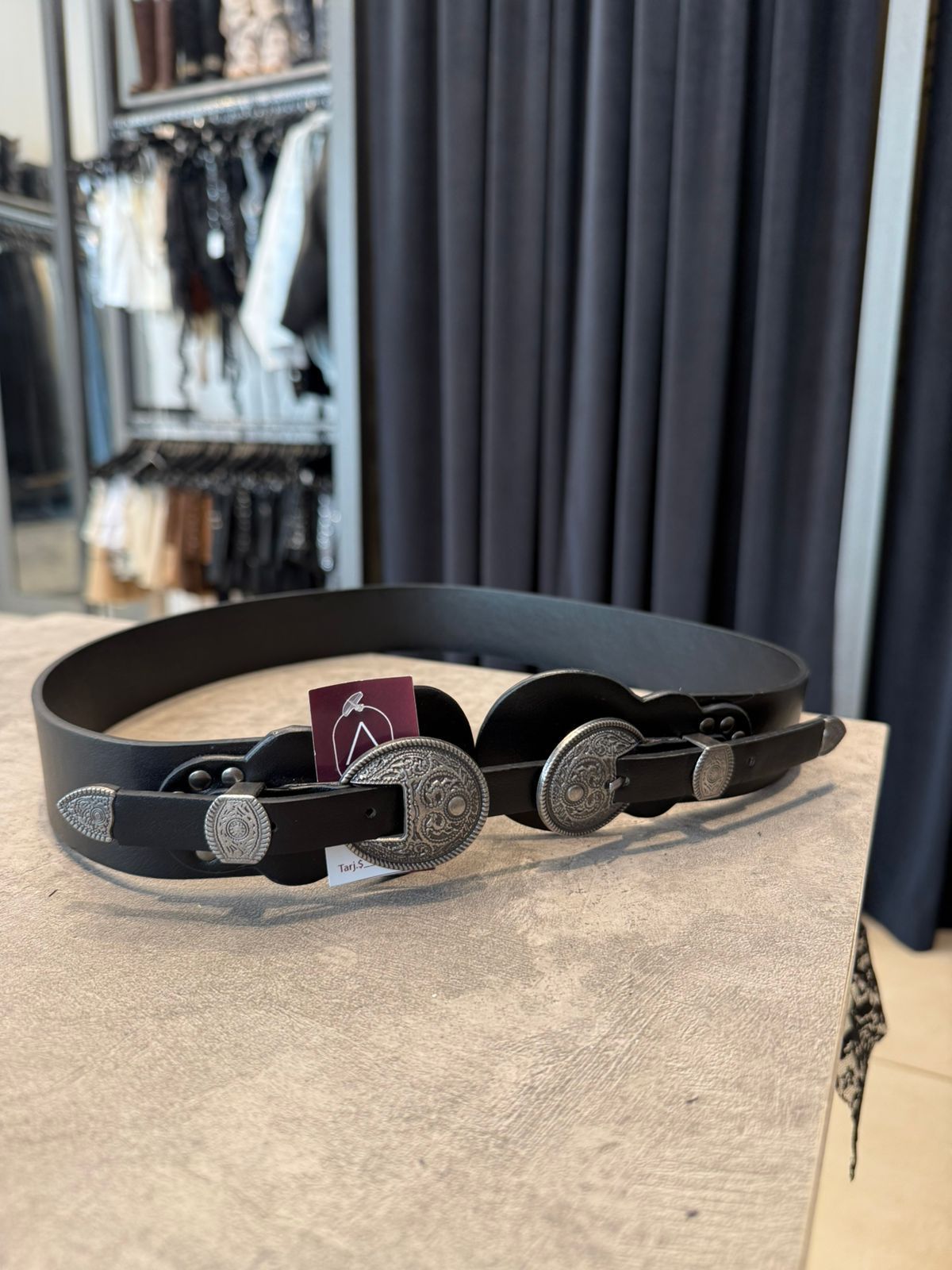 Cinturón de cuero negro con hebillas decorativas plateadas en expositor de tienda.