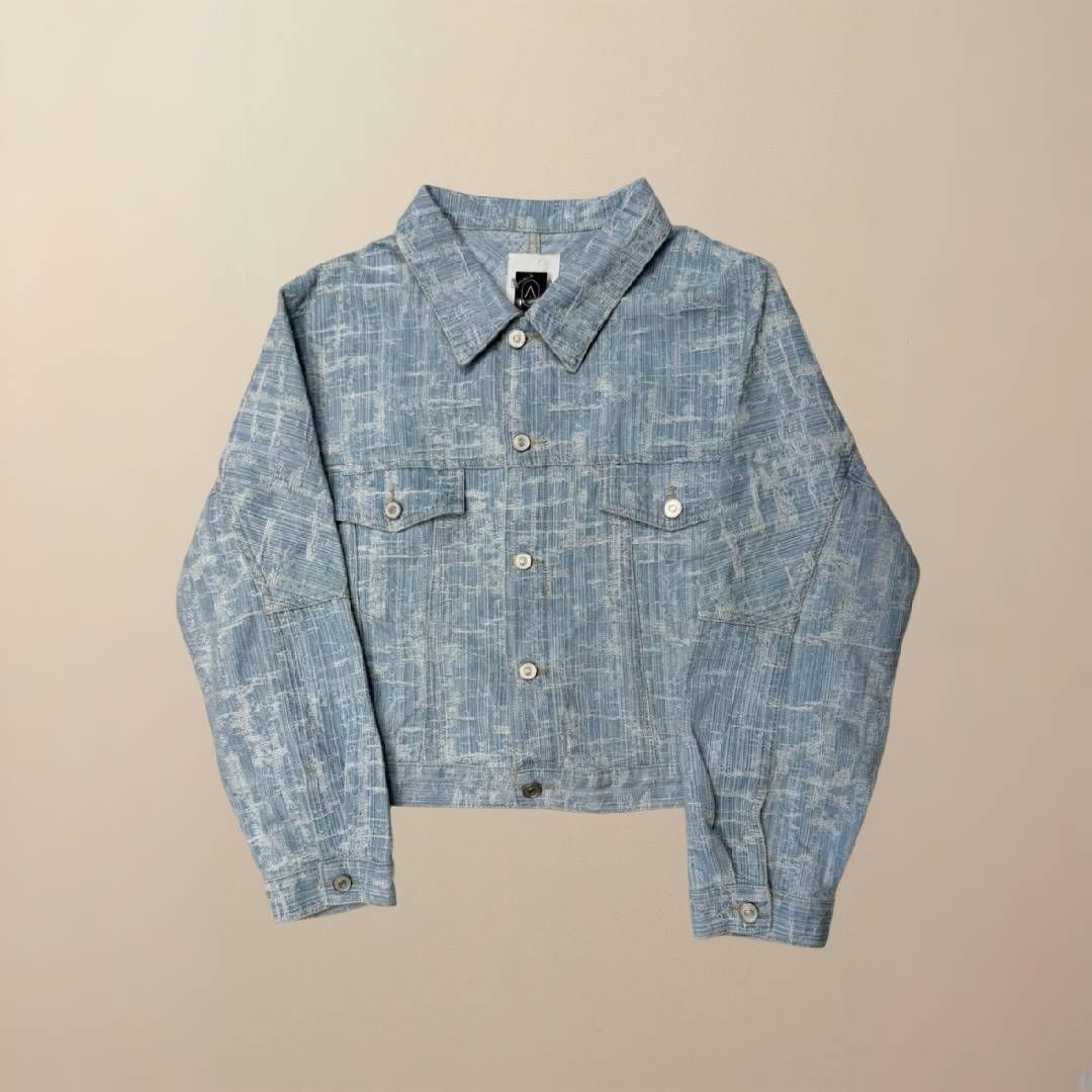Chaqueta vaquera cropped de color azul claro con estampado tipo cuadrícula sobre fondo beige.