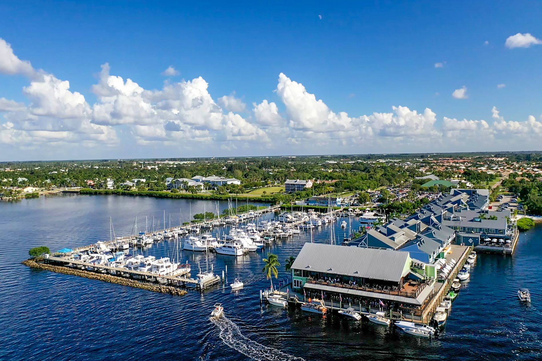 Aerial view of a marina with many docked boats. Restaurant and buildings on the right, blue water, green trees, and a partly cloudy sky. Punta Gorda, FL