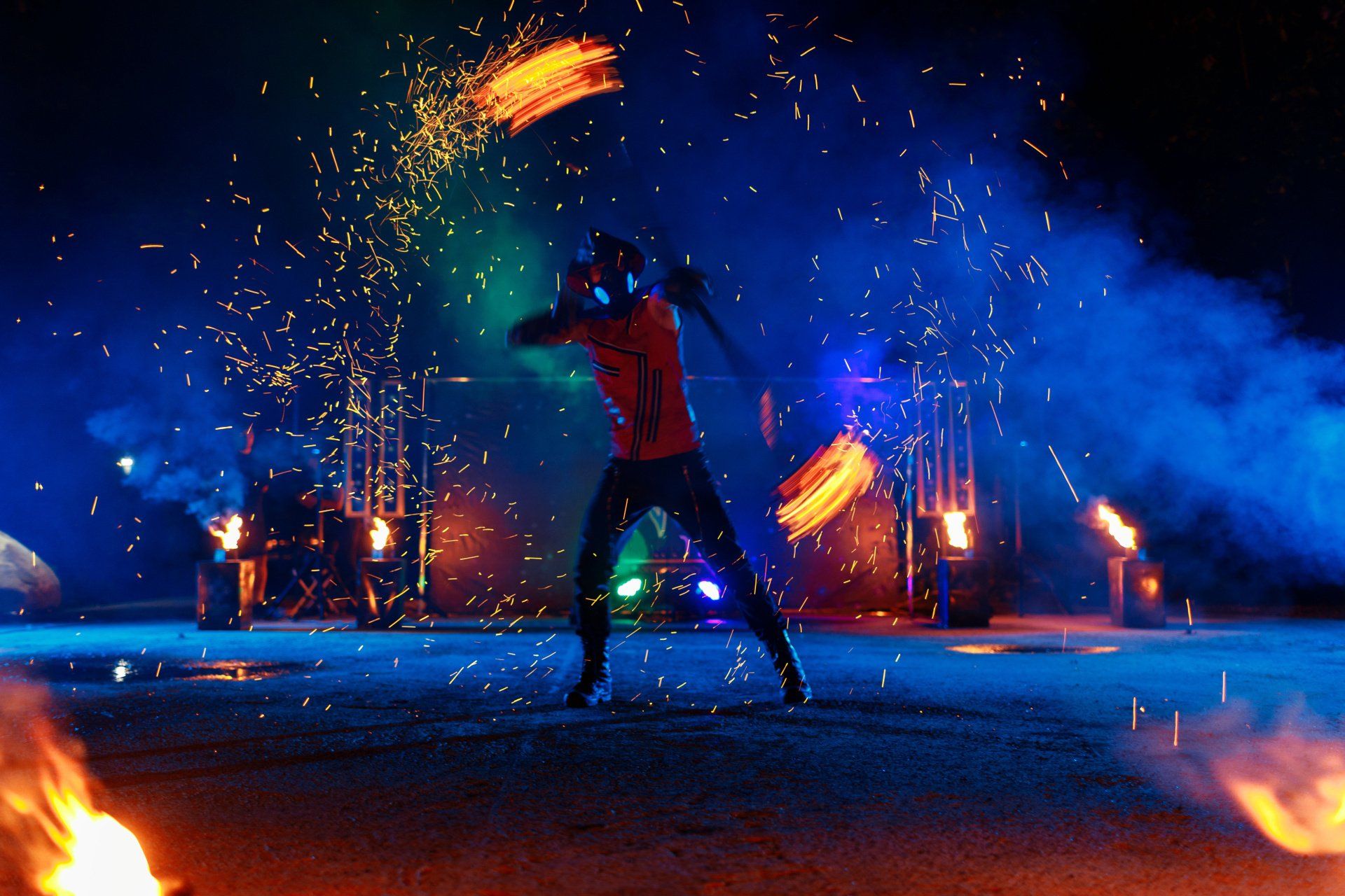 Fire performer twirls flaming baton at night with sparks and colorful lights.
