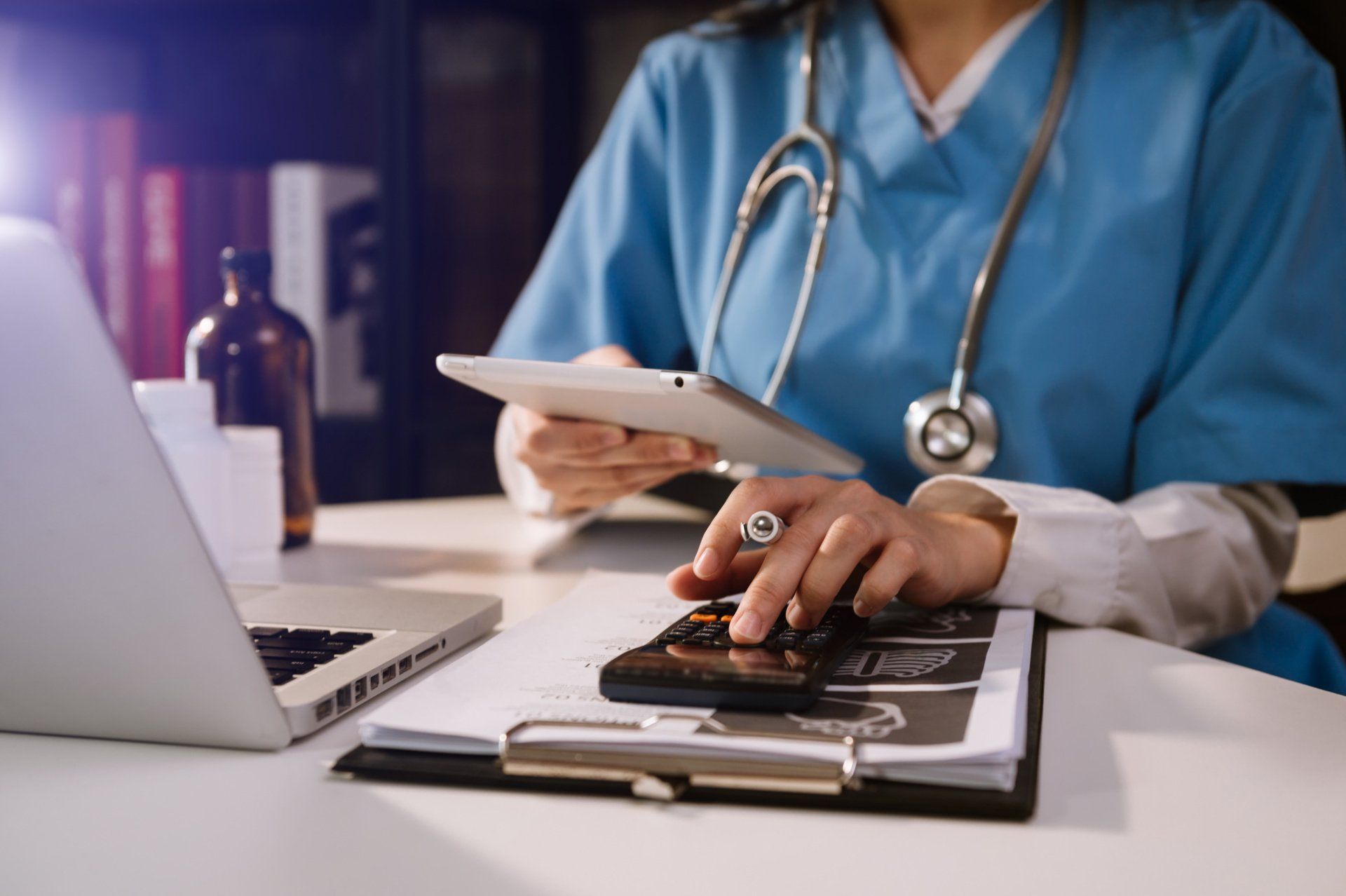 Doctor in blue scrubs using a calculator, tablet, and laptop at a desk with medical documents.