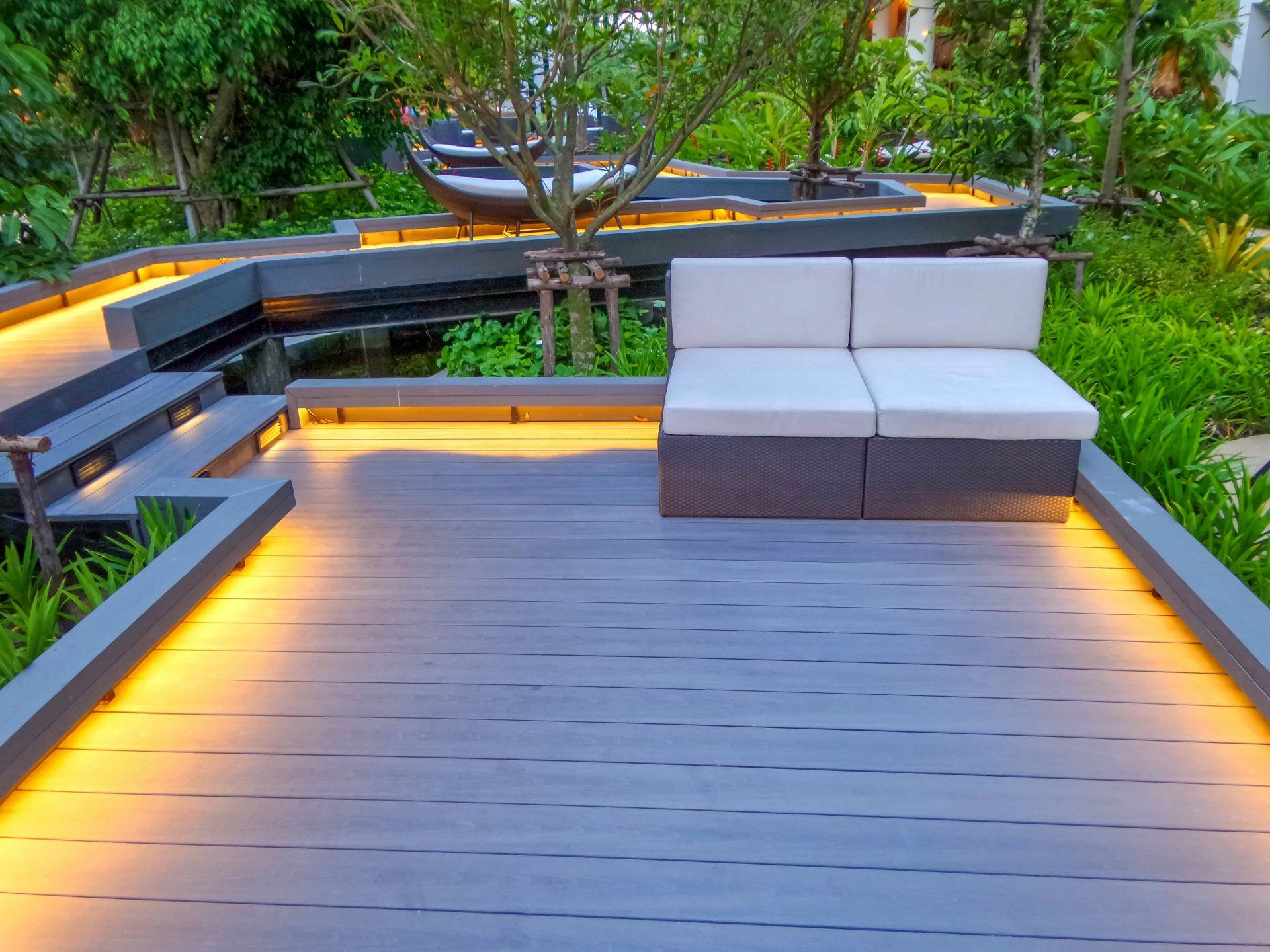 Outdoor seating area with gray wooden deck, built-in lights, and two-seat sofa. Pond and lush greenery in the background.