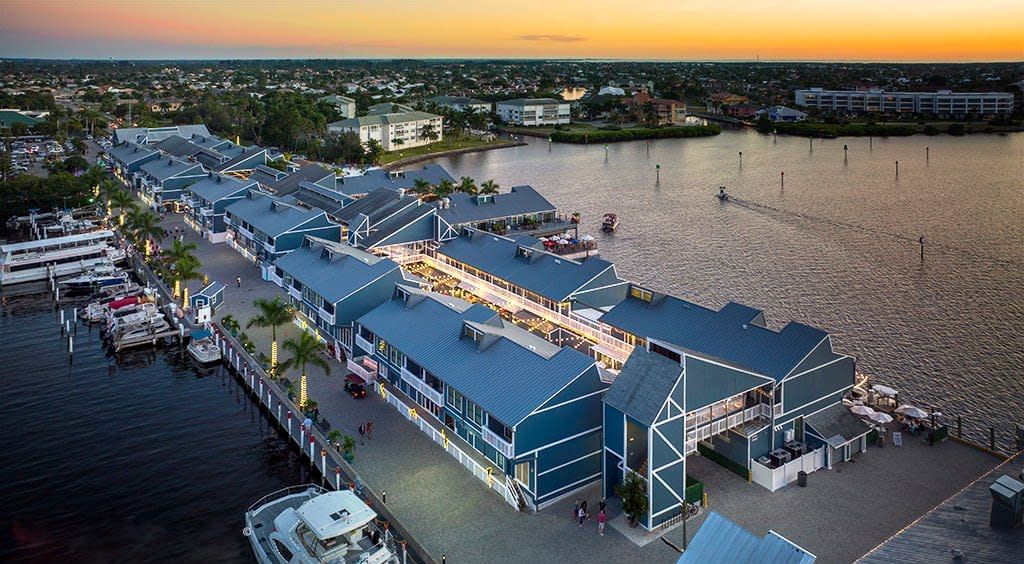 Waterfront shops with blue roofs at dusk. Boats docked, city in background.