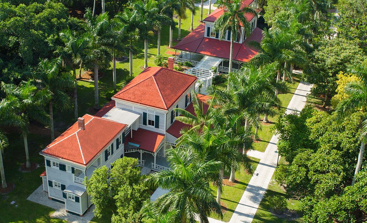 Two-story white house with red roof, surrounded by palm trees and lush greenery. A walkway leads to the house.
